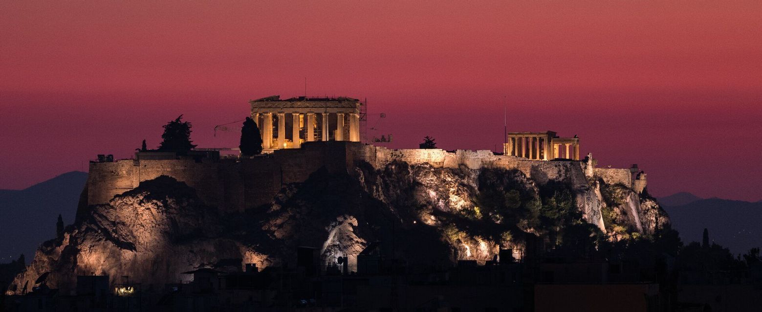 Due antichi templi con colonne sono illuminati su una collina rocciosa contro un cielo rosso intenso al tramonto.