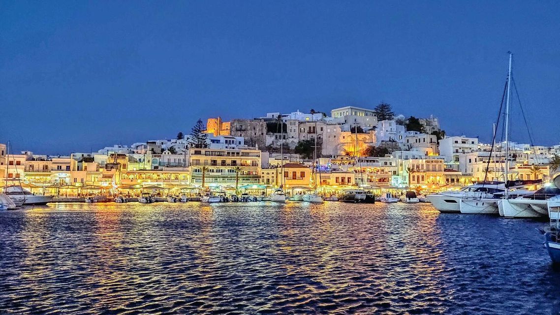 Vista dall'acqua di una città costiera illuminata al tramonto, con le luci che si riflettono sul porto dove sono ormeggiate le barche.