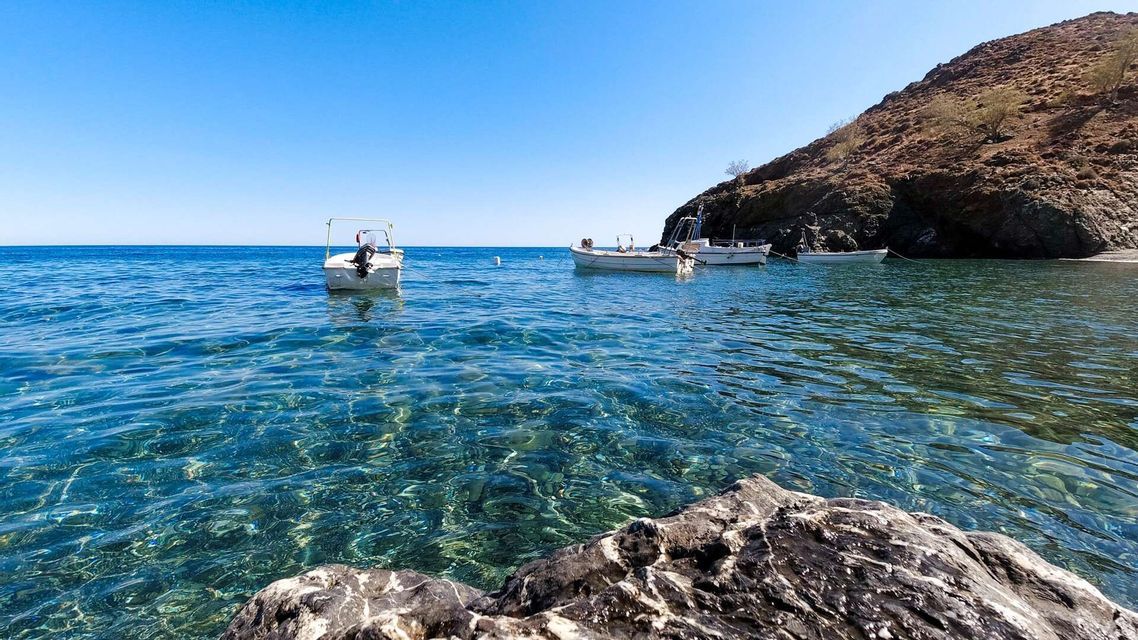 Piccole motobarche sono ormeggiate in una caletta con acqua limpida e turchese accanto a una costa rocciosa sotto un cielo azzurro brillante.