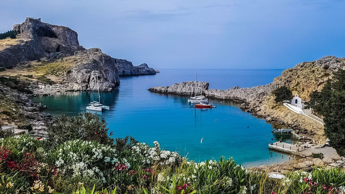 Una vista panoramica di una baia turchese con barche, scogliere rocciose e una piccola cappella bianca, incorniciata da fiori colorati.