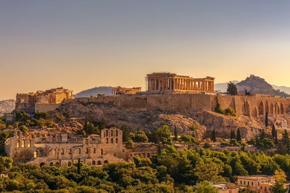 L'antico tempio del Partenone sulla collina dell'Acropoli, illuminato dalla luce dorata del tramonto, con rovine e alberi verdi sottostanti.