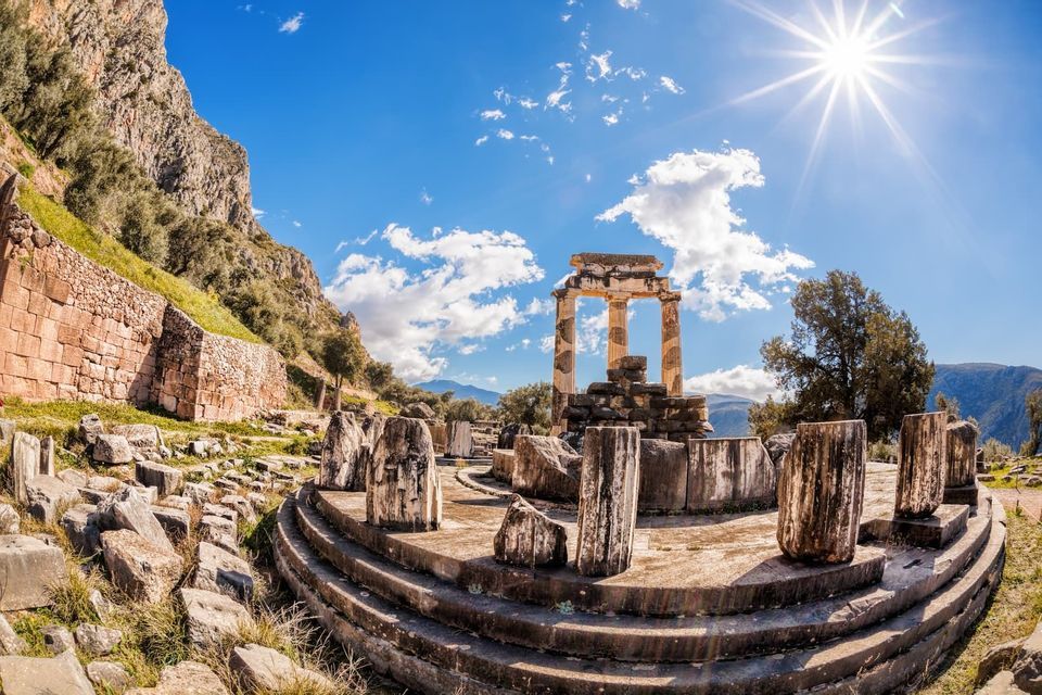 Le rovine di un antico tempio circolare con colonne di pietra su un fianco di montagna sotto un sole splendente e cielo azzurro.