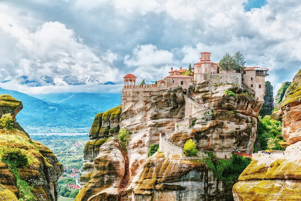 Un monastero di pietra eretto su uno sperone di roccia a picco, con una scalinata tortuosa e una vista su una vasta valle di montagna sotto un cielo nuvoloso.
