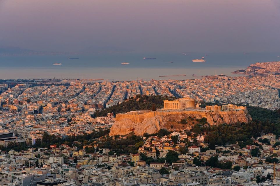 Antiche rovine su una collina brillano al tramonto, affacciate su un denso panorama urbano che si estende fino al mare.