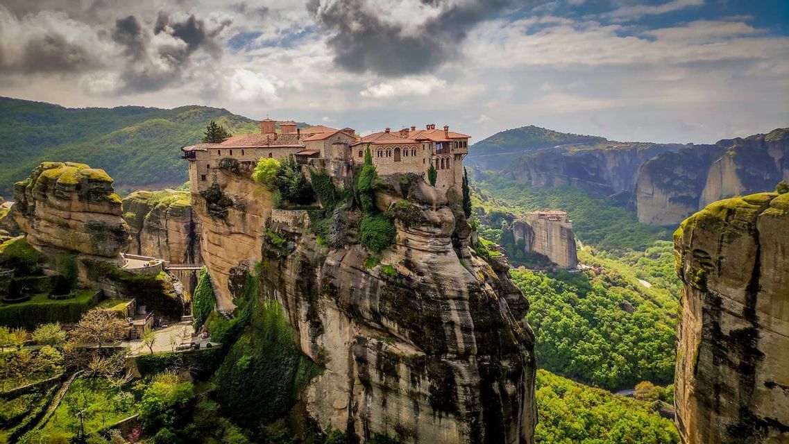 Un monastero di pietra con tetto in tegole rosse sorge su un'alta e ripida colonna di roccia, dominando una vasta valle verde e montuosa.