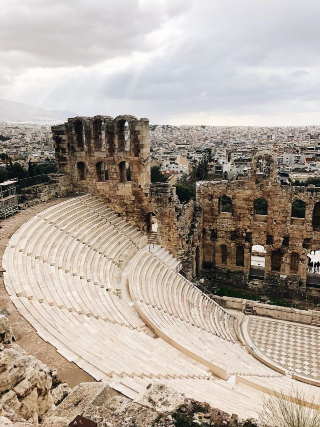 Un antico anfiteatro in pietra con gradinate si affaccia su un denso paesaggio urbano sotto un cielo nuvoloso.