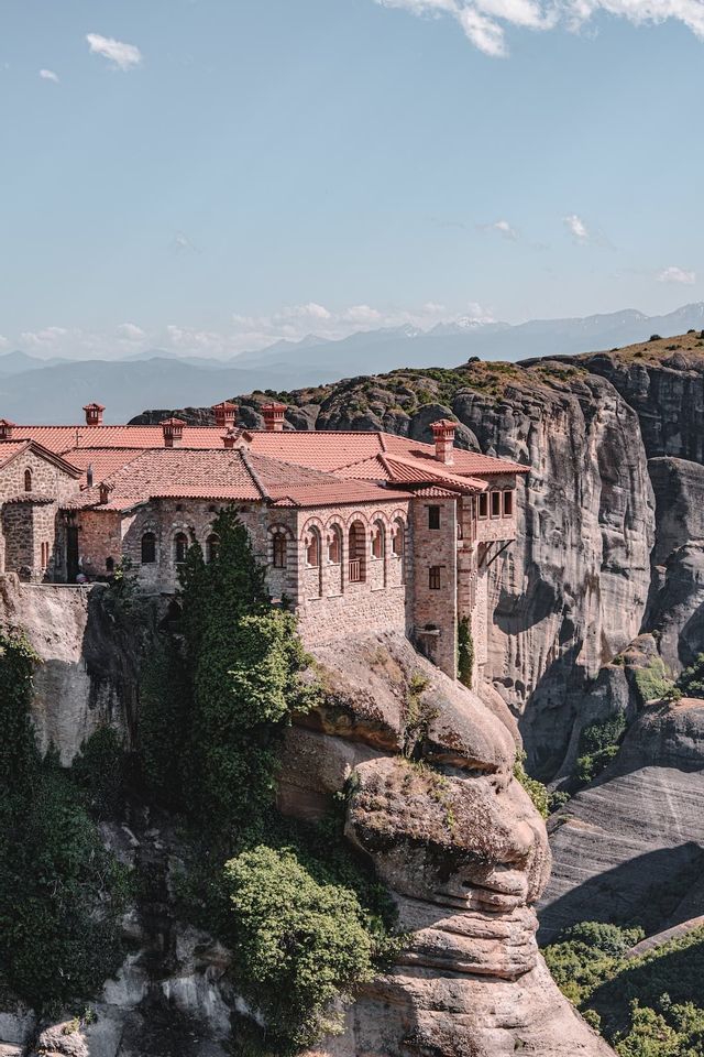 Un monastero di pietra con tetto in tegole rosse costruito sul bordo di una ripida scogliera rocciosa, con montagne distanti sullo sfondo.