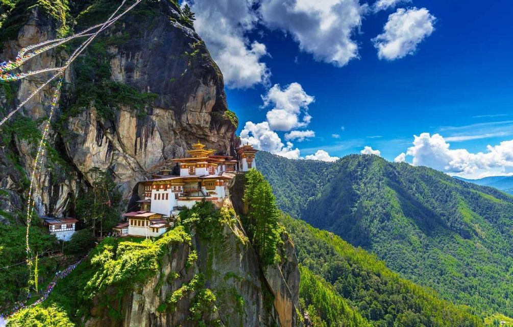 Un complesso monastico con pareti bianche e tetti dorati è costruito in una scogliera a picco che domina una valle di verdi montagne boscose.
