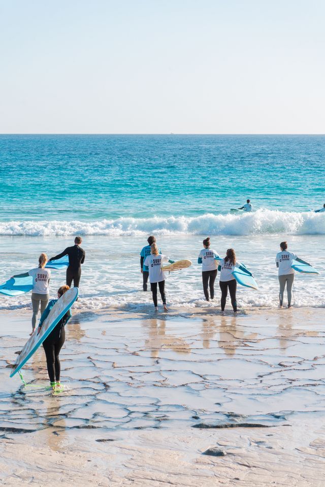 Un gruppo WeRoad, in muta da sub, porta tavole da surf nell'oceano da una costa rocciosa a terrazze per una lezione di surf.