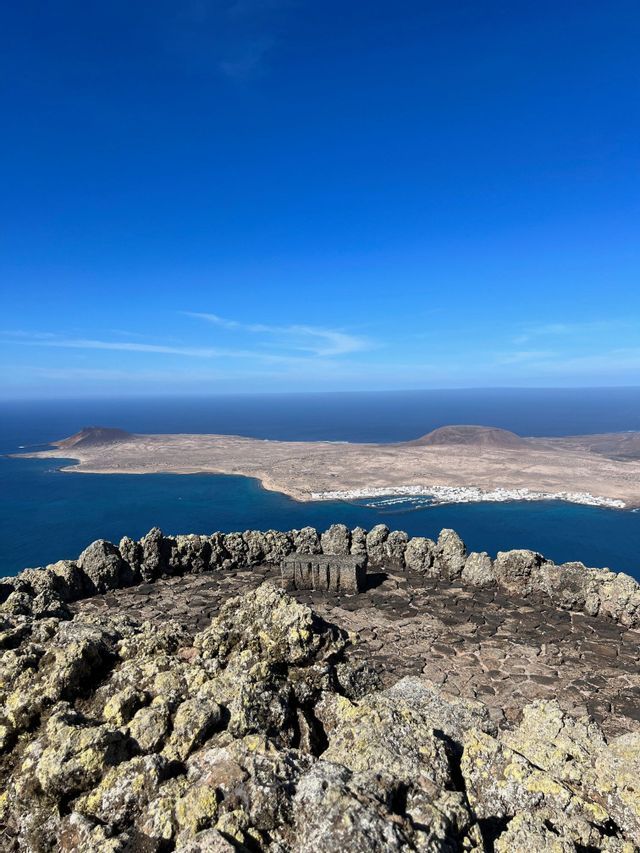 Un punto panoramico in pietra su una scogliera domina un'isola vulcanica con un piccolo paese costiero, circondata da un mare blu profondo.