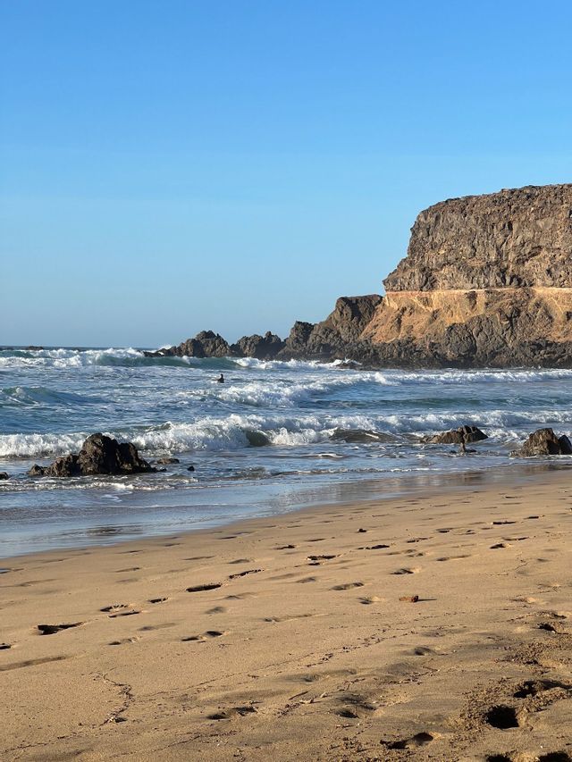 Una spiaggia sabbiosa con impronte porta all'oceano, dove una persona attende tra le onde vicino a una scogliera rocciosa sotto un cielo blu chiaro.