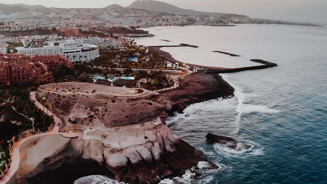 Una vista aerea di una città costiera al tramonto, con edifici lungo una costa rocciosa e spiagge di sabbia scura accanto all'oceano.