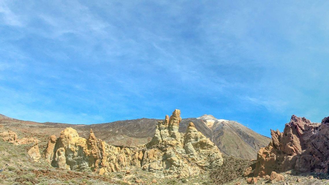 Formazioni rocciose vulcaniche frastagliate si ergono da un paesaggio aspro, con una lontana cima montuosa visibile sotto un vasto cielo blu.