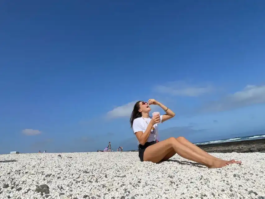 Una donna con occhiali da sole è seduta su una spiaggia di ciottoli bianchi, mentre mangia un pezzo di frutta da una bevanda rosa sotto un cielo azzurro e limpido.