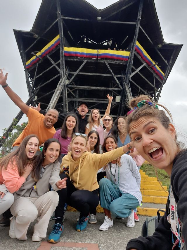 Un viaggio di gruppo WeRoad si fa un selfie energico sui gradini davanti a un'alta torre di osservazione con una bandiera colombiana.