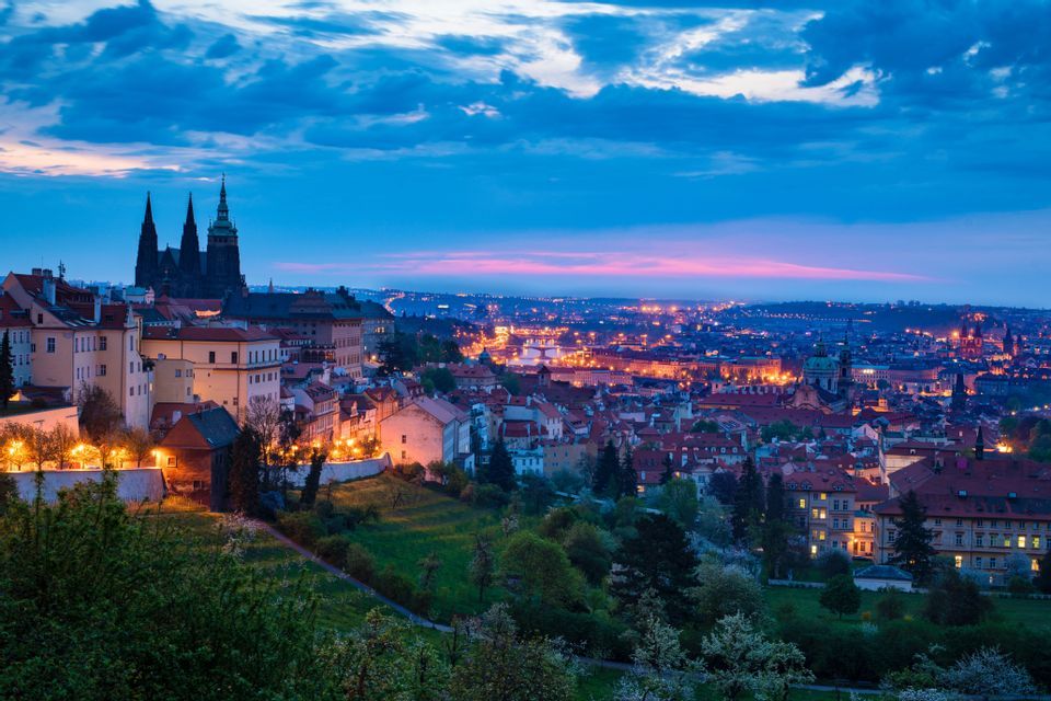 Un horizonte urbano con una catedral gótica silueteada al anochecer, vista desde una colina con edificios iluminados debajo.
