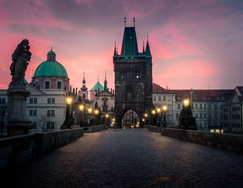 Un puente histórico de adoquines con estatuas y farolas encendidas al anochecer, con una torre de piedra y edificios abovedados bajo un cielo rosado.