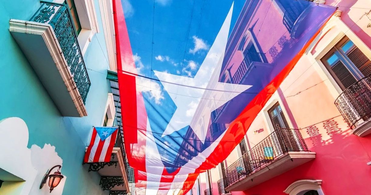 Una grande bandiera portoricana si estende su una stretta strada tra edifici colorati con balconi sotto un cielo blu.
