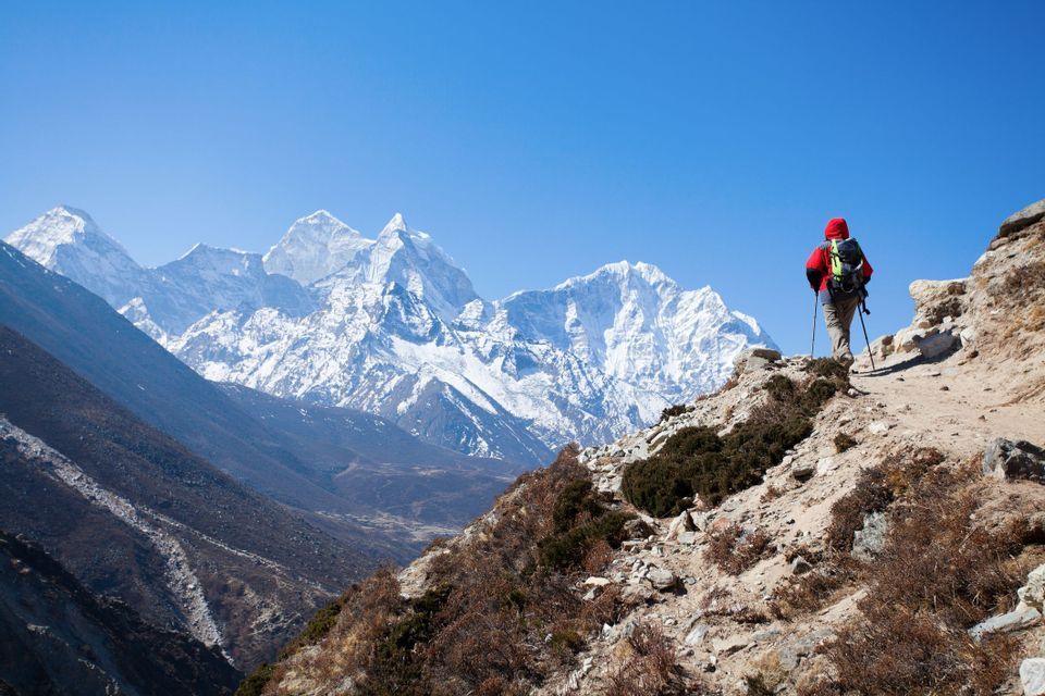 A hiker with a backpack and poles stands on a rocky trail, gazing out at a vast snow-capped mountain range under a blue sky.