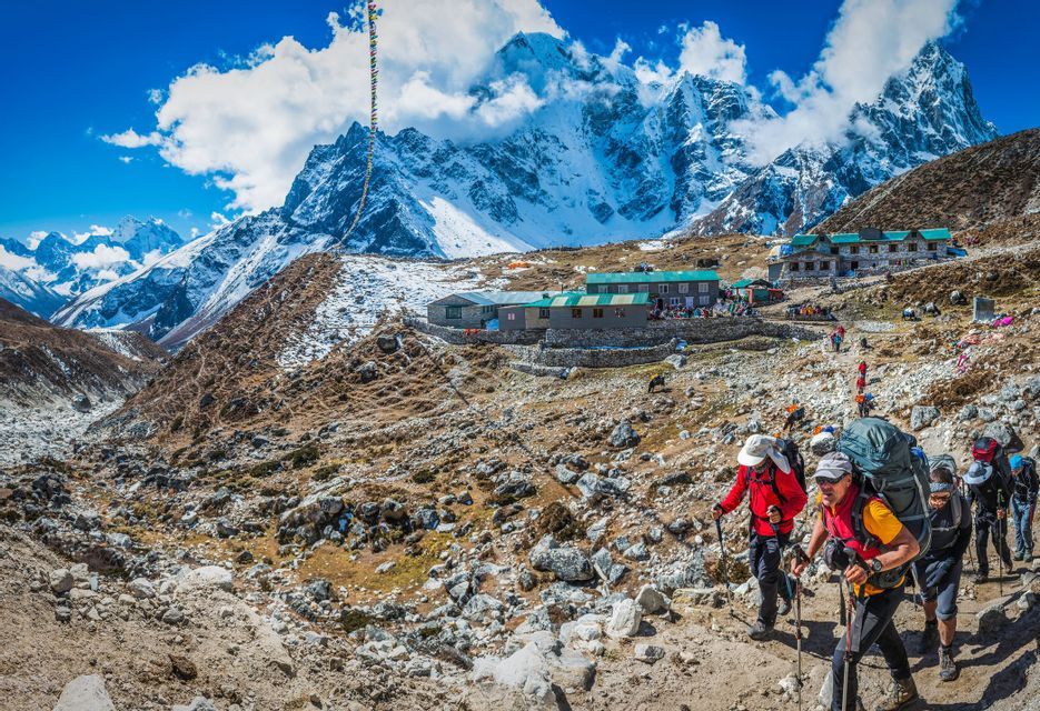 Un voyage de groupe WeRoad en randonnée sur un sentier rocheux menant à un refuge de montagne, avec de grands sommets enneigés en arrière-plan.