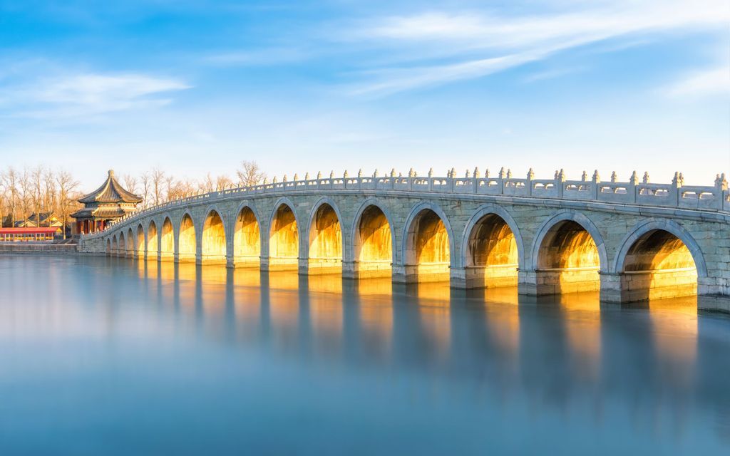Un lungo ponte di pietra a più arcate attraversa acque calme, i suoi archi risplendono di luce solare dorata sotto un cielo azzurro.