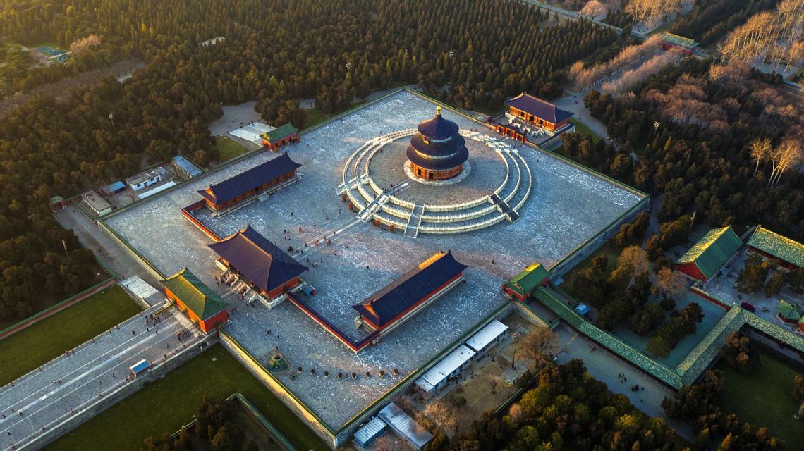 Una veduta aerea di un tradizionale complesso di templi cinesi con una pagoda circolare centrale, circondato da un ampio cortile e una fitta foresta al tramonto.