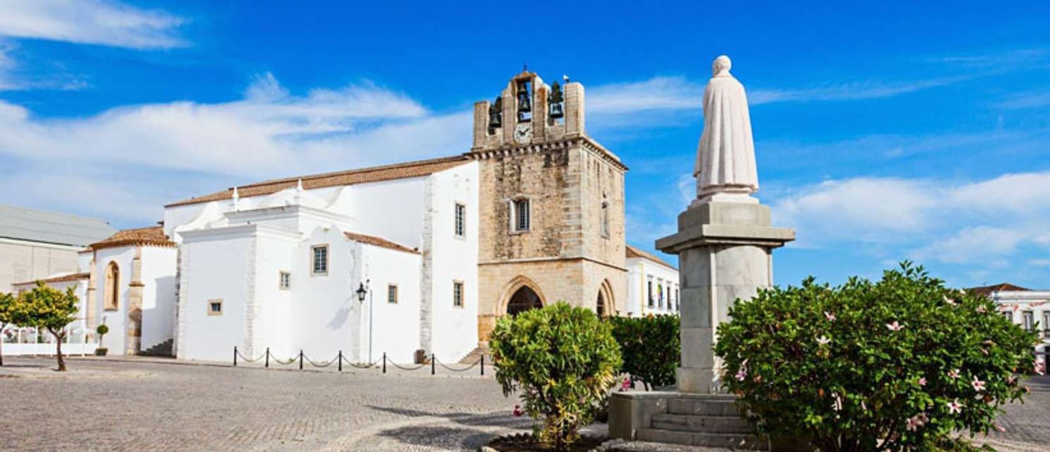 Una chiesa imbiancata a calce con un campanile in pietra e una statua si ergono in una piazza acciottolata sotto un cielo azzurro brillante.