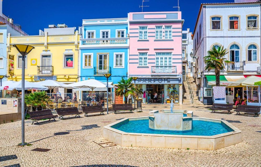 Una fontana di pietra si trova in una piazza acciottolata costeggiata da edifici colorati, caffè e palme sotto un cielo azzurro limpido.