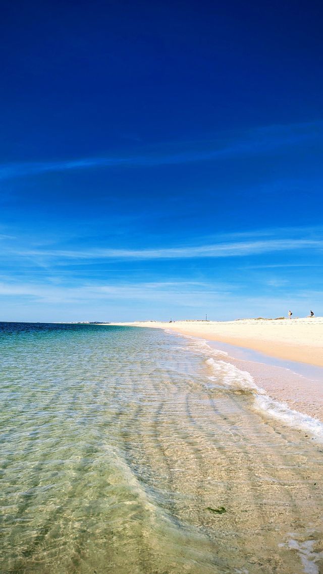Acqua limpida e bassa increspa una spiaggia ampia e sabbiosa sotto un vasto cielo blu intenso.