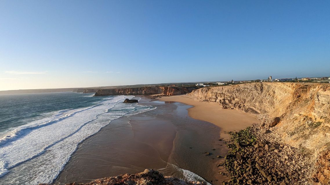 Un'ampia vista di una spiaggia sabbiosa incastonata tra scogliere rocciose, con le onde dell'oceano che si infrangono a riva sotto un cielo azzurro e limpido.