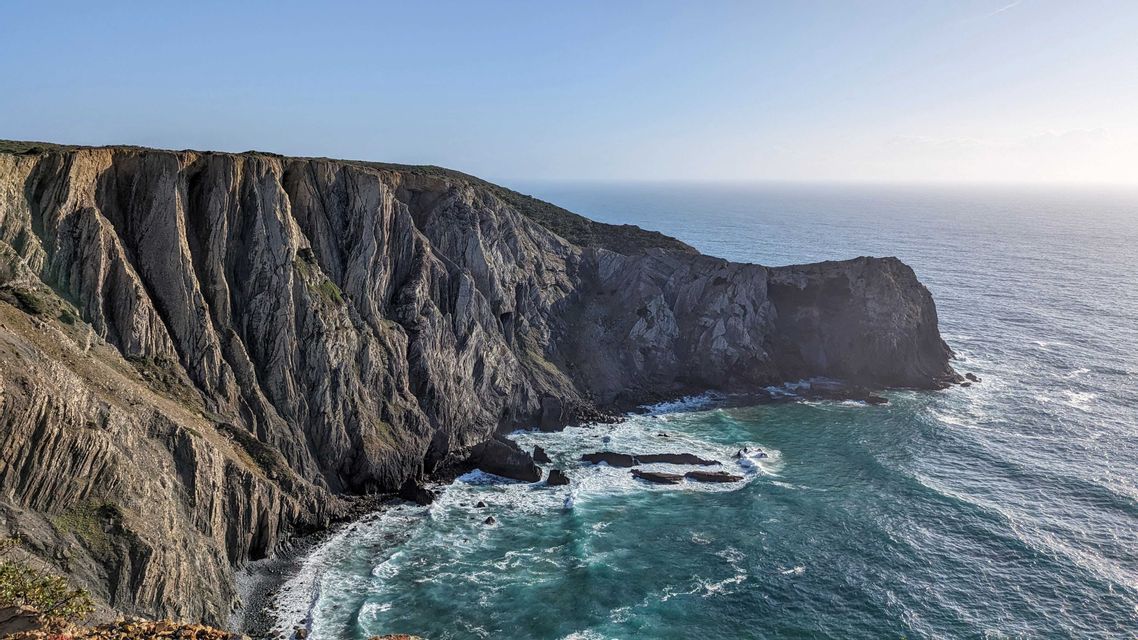 Scogliere rocciose a strati a picco incontrano un oceano turchese, con onde che si infrangono contro la costa rocciosa.