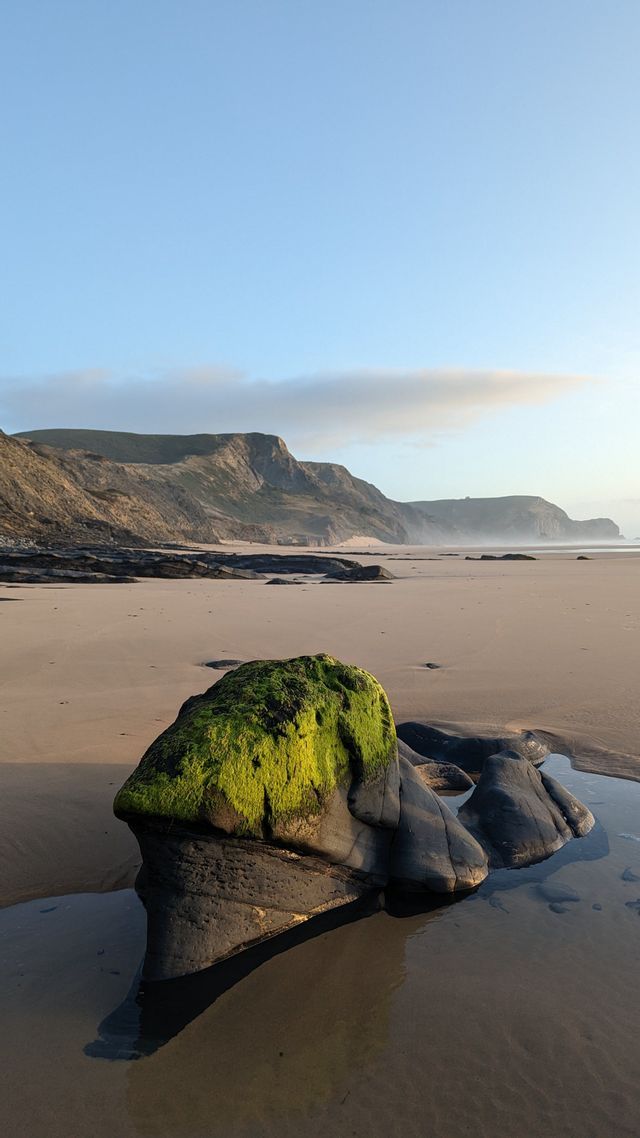 Una grande roccia coperta di muschio verde si trova su una spiaggia sabbiosa con scogliere sullo sfondo sotto un cielo sereno.