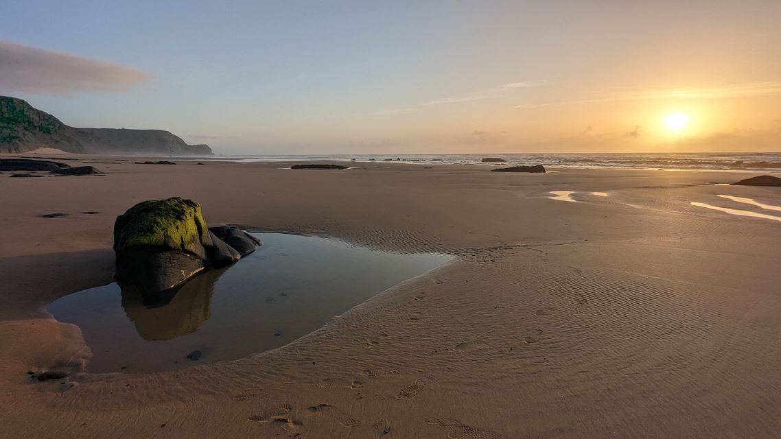 Un'alba dorata su un'ampia spiaggia sabbiosa, con una roccia muschiosa e una pozza di marea riflettente in primo piano.