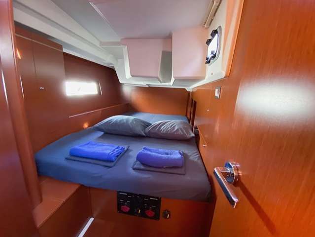 Un letto matrimoniale ben fatto con lenzuola blu all'interno di una cabina barca compatta con pareti rivestite in legno e un piccolo oblò.