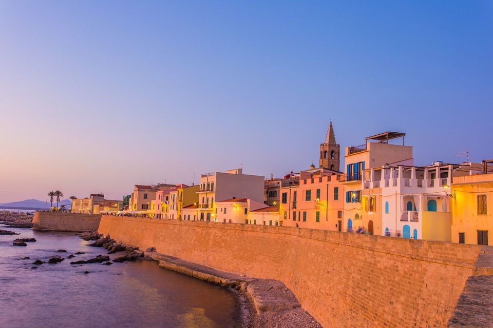 Le mura fortificate di una città costiera con case colorate sono illuminate dalla calda luce del tramonto.