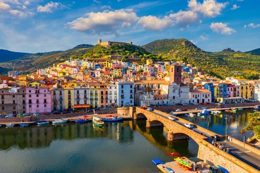 Un colorato borgo collinare con castello si riflette nel fiume, attraversato da un ponte in pietra ad arco.
