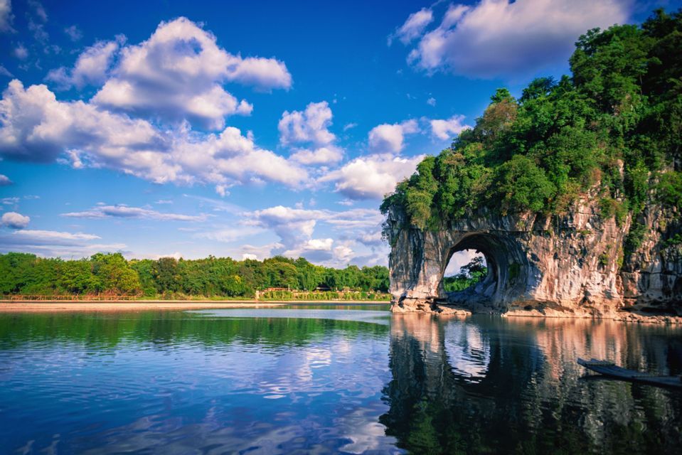 Un'ampia formazione rocciosa con un arco naturale si erge in un fiume calmo, riflettendosi sull'acqua sotto un cielo azzurro nuvoloso.