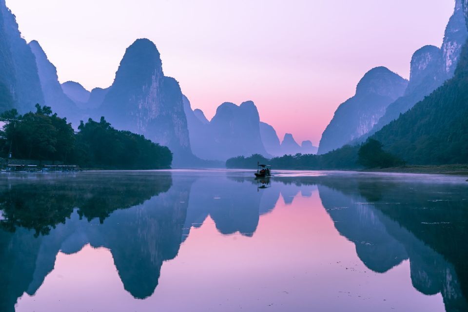 Una barca naviga su un fiume calmo, riflettendo montagne carsiche in controluce contro un cielo viola al tramonto.