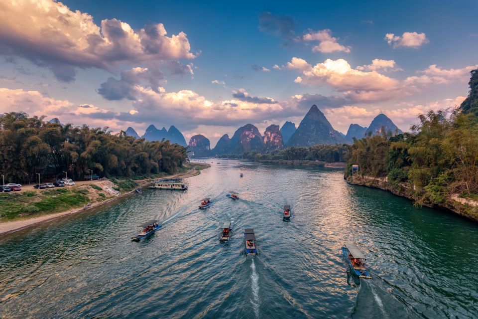 Barche turistiche navigano su un ampio fiume, circondato da foreste verdi e imponenti montagne carsiche, sotto un cielo parzialmente nuvoloso.