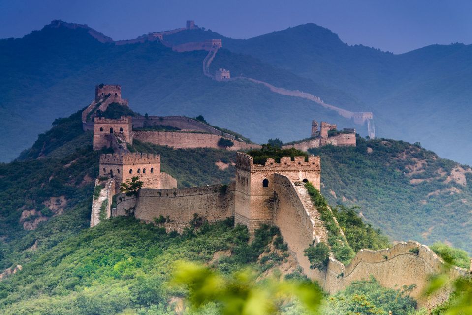 La Grande Muraglia Cinese si snoda tra montagne verdi e lussureggianti, con torri di guardia che si estendono verso uno sfondo blu e sfumato.