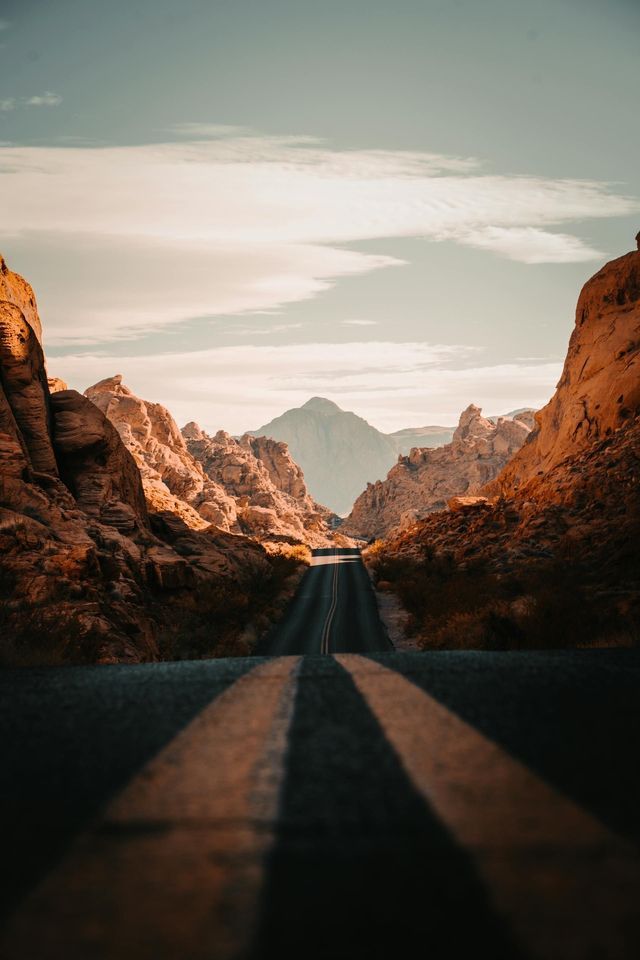 Panoramica dal basso di una strada asfaltata con linee gialle che attraversa un canyon di formazioni rocciose arancioni verso una montagna distante.