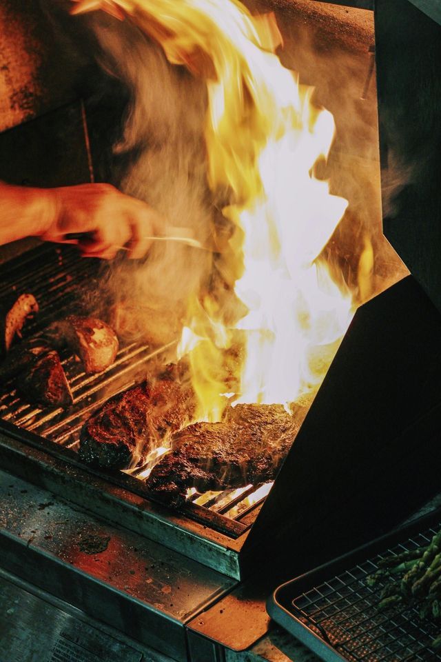 Una mano con le pinze gira le bistecche su una griglia per barbecue mentre grandi fiamme divampano.