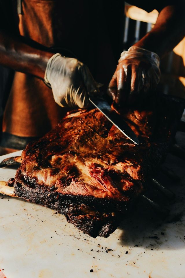 Una persona con guanti affetta un grosso taglio di carne affumicata con un coltello su un tagliere bianco.