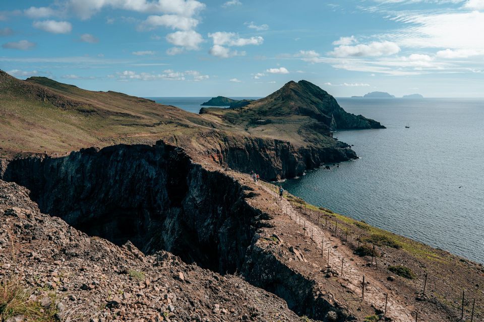 Un viaje en grupo de WeRoad haciendo senderismo por un sendero estrecho a lo largo de un acantilado costero rocoso, con el océano e islas distantes a la vista.