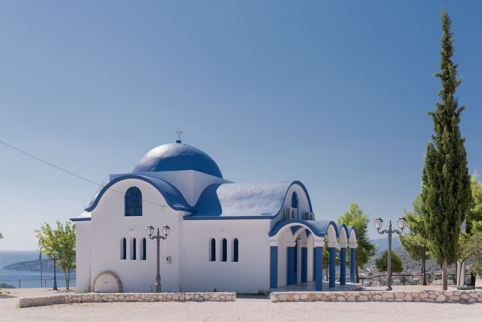 Una chiesa bianca con una cupola blu e accenti sul tetto si erge su una collina che domina il mare sotto un cielo limpido.