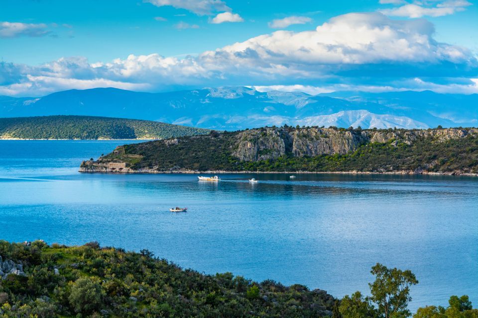 Piccole barche su un mare calmo e blu con una costa verde e rocciosa e montagne distanti sotto un cielo parzialmente nuvoloso.