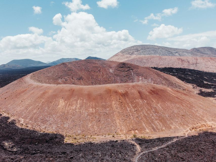 Un grande cratere vulcanico rossastro si trova all'interno di un vasto paesaggio di roccia lavica nera sotto un cielo parzialmente nuvoloso.