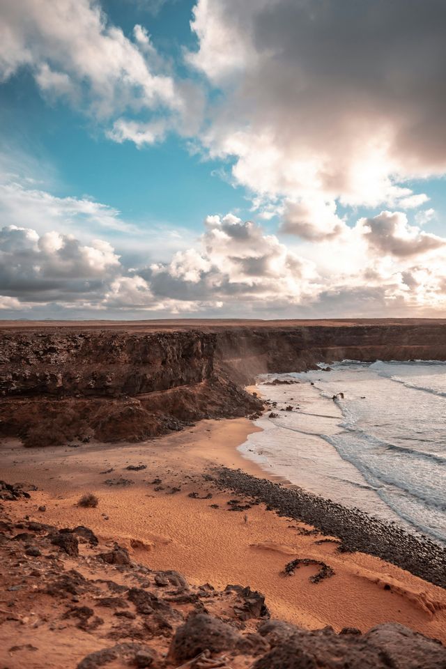 Una vista dall'alto di una costa rocciosa con sabbia arancione, scogliere scure e onde oceaniche sotto un cielo parzialmente nuvoloso.