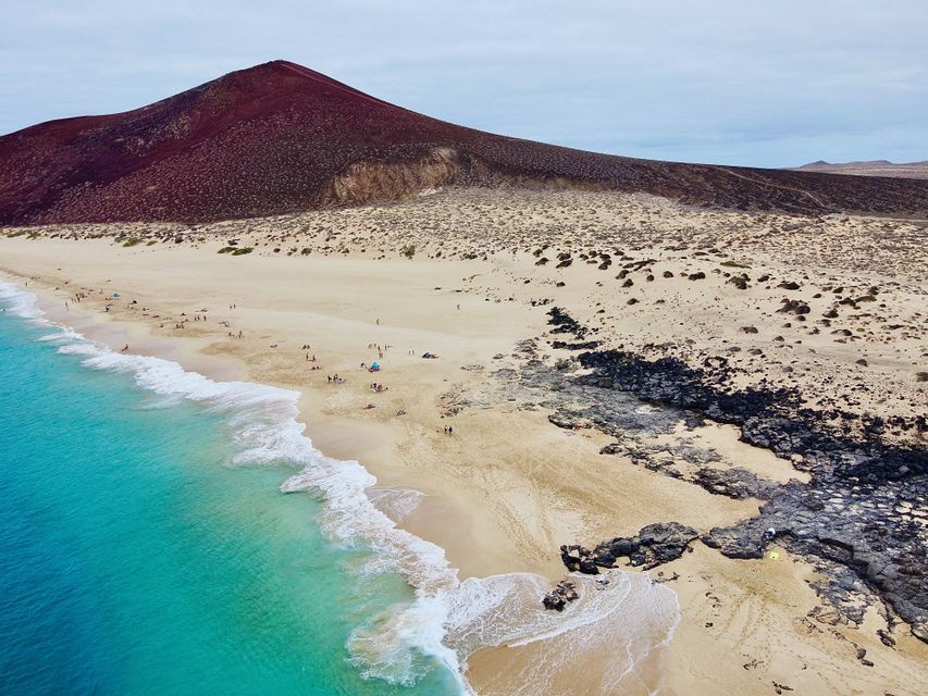 Una vista aerea di una spiaggia sabbiosa con acqua turchese e rocce vulcaniche, con una grande montagna rossa sullo sfondo.
