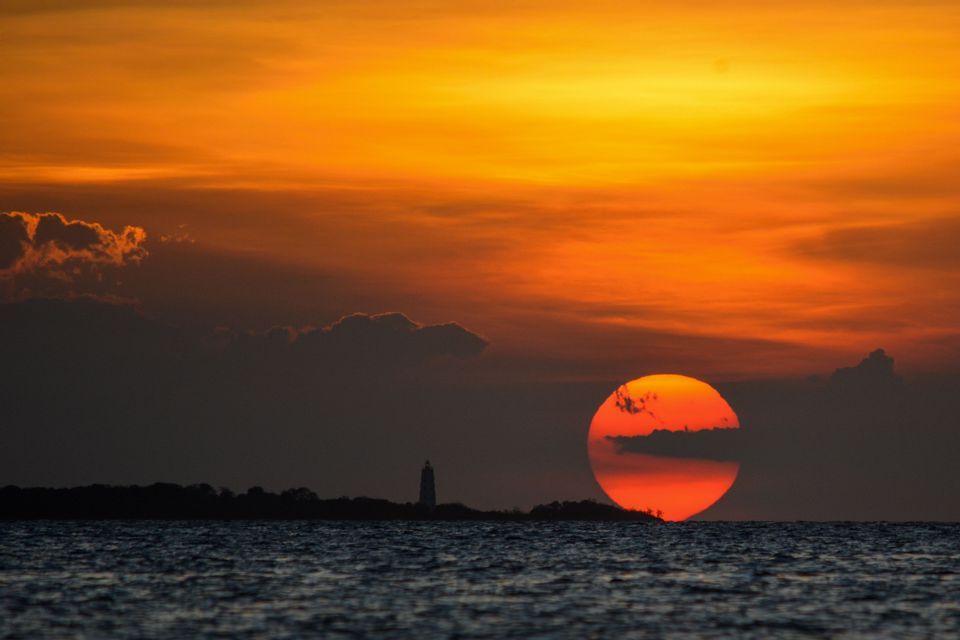 Un grande sole arancione tramonta sull'oceano con un faro stagliato all'orizzonte sotto un cielo arancione splendente.